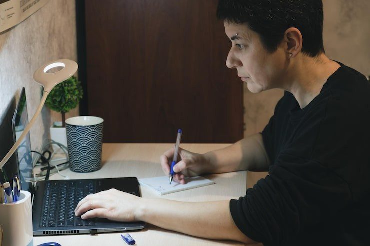 middleaged-brunette-woman-sits-desk-front-laptop-screen-makes-notes-notebook_259471-2301